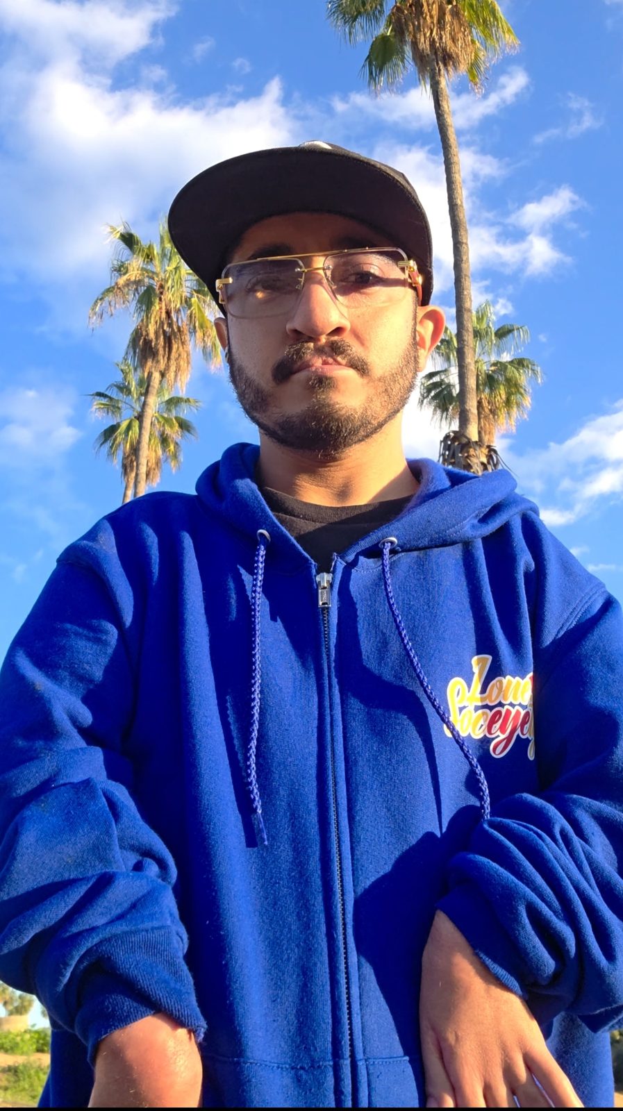 Lone.Fractal standing outdoors in front of palm trees under a blue sky, wearing a cap, gold-framed glasses and a blue zip-up hoodie